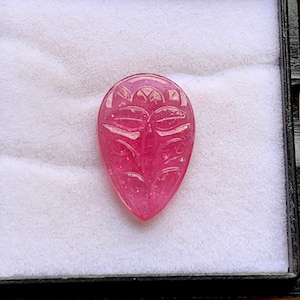 May include: A teardrop-shaped, translucent pink gemstone featuring an intricate, symmetrical floral carving on its surface. The stone has a smooth, polished finish and is displayed in a box with a white, soft-textured interior lining.