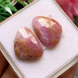 May include: Two pear-shaped faceted gemstones with a pink and orange colour palette. The stones feature a polished, multi-faceted surface that reflects light, showcasing natural internal inclusions and variations in the stone's colour and texture.