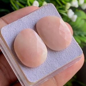 May include: Two oval, pale pink gemstones with faceted surfaces. They are presented on a light grey, rectangular surface, possibly for display or storage. The background is blurred, focusing attention on the gemstones.