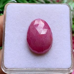 May include: A faceted, pear-shaped gemstone with a deep pinkish-red hue. The stone features a polished surface with multiple geometric facets that reflect light, and it is centered within a square, white, foam-lined display box.
