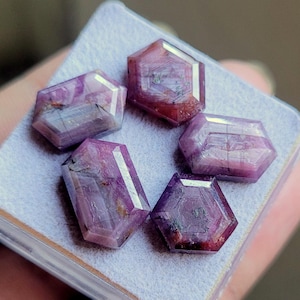 May include: Five faceted, hexagonal gemstones in shades of purple and red. The gems are displayed on a light grey surface. The gemstones have a polished, reflective surface, with visible internal patterns and inclusions. The overall aesthetic is one of natural beauty and craftsmanship.