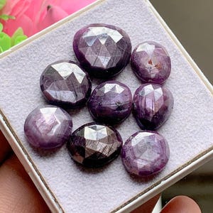 May include: A collection of seven faceted, round, deep purple and magenta gemstones arranged on a light grey display pad. Each stone features a polished, multi-faceted surface that reflects light, showcasing varying shades of dark violet and reddish-purple hues.
