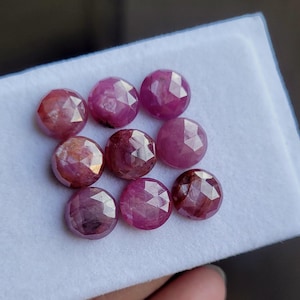 May include: Nine faceted, round gemstones in shades of pink and red are displayed on a white surface. The gems have a polished, reflective surface and are arranged in a grid pattern. The gemstones are likely rubies.