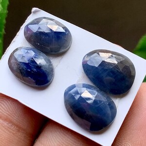 May include: Four oval-shaped, faceted blue sapphire gemstones arranged on a white rectangular card. Each stone features a polished, multi-faceted surface that reflects light, displaying deep navy and royal blue hues with natural internal inclusions.