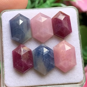 Op de afbeelding: Zes gefacetteerde zeshoekige edelstenen gerangschikt in twee rijen in een witte displaydoos. De stenen hebben verschillende kleuren, waaronder diepblauw, lichtroze en donker magenta, elk met een gepolijst oppervlak dat het licht geometrisch weerkaatst.