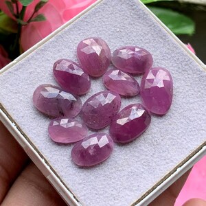 Puede incluir: Una colección de nueve rubíes rosas facetados de forma ovalada dispuestos sobre una base de fieltro gris claro. Cada piedra preciosa presenta una superficie pulida con múltiples facetas que refleja la luz, mostrando tonos de magenta y rosa intenso.