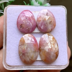 May include: Four faceted, oval-shaped pink and white gemstones arranged in a square formation inside a clear plastic display case. The stones exhibit a mix of opaque and translucent textures with natural inclusions and a polished, reflective surface.