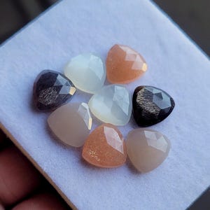 Puede incluir: Una colección de ocho gemas triangulares facetadas en varios colores, incluyendo melocotón, gris y blanco. Las gemas están dispuestas sobre una superficie blanca, mostrando sus superficies pulidas y variaciones de color únicas. Ideales para la fabricación de joyas.
