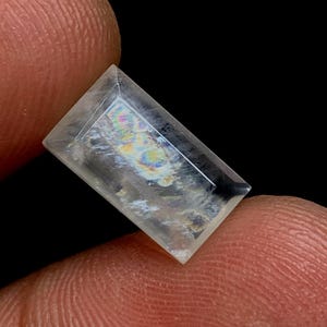 以下が含まれることがあります： 虹色のイリデッセンスを持つ透明な長方形の宝石。 宝石は黒い背景に対して2本の指の間に保持されています。 宝石はカットされたエッジを持つ長方形の形状をしています。