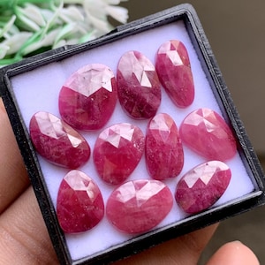 May include: A collection of nine faceted, deep pink, oval and pear-shaped gemstones arranged inside a small, square black display box with a white foam insert. Each stone features a polished, multi-faceted surface that reflects light.