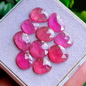 Può includere: Una collezione di nove gemme rosa di forma ovale e sfaccettate, disposte a grappolo. Ogni pietra presenta una superficie lucida a facce multiple che riflette la luce, mostrando una tonalità magenta vivace e traslucida su tutto il set.