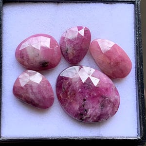 May include: Five faceted pink ruby gemstones of varying shapes and sizes arranged on a white display pad. Each stone features a polished surface with visible natural inclusions, dark mineral flecks, and a translucent to opaque pink colour profile.
