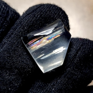 May include: A clear, faceted gemstone with internal rainbow iridescence. The gem is held against a dark, textured background, highlighting its polished surfaces and light refraction. The shape is a modified trapezoid.