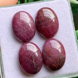 May include: Four oval-shaped, polished ruby gemstones with a deep magenta-red hue. Each stone features a smooth, rounded cabochon cut with visible natural internal inclusions and a glossy surface, arranged in a two-by-two grid pattern.