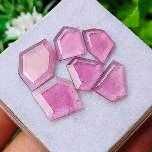 Può includere: Una collezione di sei pietre preziose rosa sfaccettate a forma di scudo pentagonale. Ogni pietra presenta una superficie lucida con bordi geometrici distinti e una tonalità rosa traslucida, disposte su un supporto espositivo grigio chiaro.