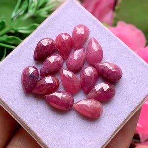 Peut inclure: Un ensemble de treize rubis facettés en forme de poire, d'une teinte magenta-rose profond. Chaque pierre précieuse présente une surface polie à facettes multiples qui reflète la lumière, disposée en grappe sur un support carré en velours violet clair.