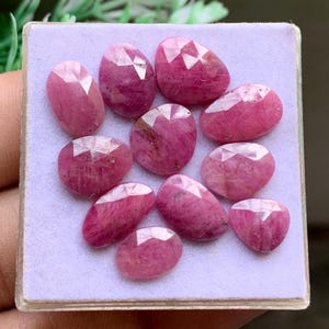 May include: A collection of ten faceted, oval and pear-shaped pink rubies arranged on a square, light purple display pad. Each gemstone features a polished surface with geometric facets that reflect light, showcasing deep magenta and pink hues.
