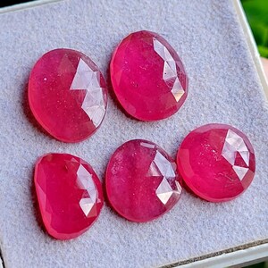 Peut inclure: Cinq pierres précieuses rose vif, de forme ovale et à facettes, disposées sur une surface texturée gris clair. Chaque pierre présente une surface polie à facettes multiples qui reflète la lumière, révélant une teinte magenta saturée.