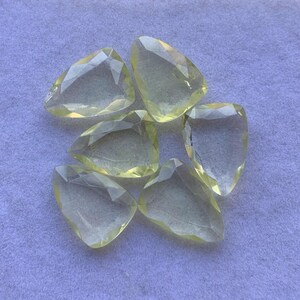 May include: Six faceted, pear-shaped gemstones in a pale yellow colour. The gems are arranged on a light purple surface, each displaying multiple facets that catch the light. The stones vary slightly in size and shape.
