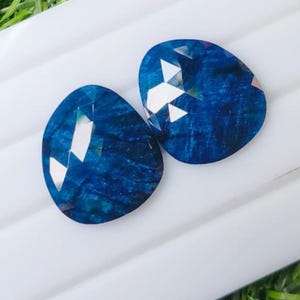 Puede incluir: Dos piedras preciosas facetadas en forma de lágrima de un color azul profundo y vibrante. Las piedras presentan una superficie pulida con texturas internas visibles y facetas geométricas nítidas que reflejan la luz con brillos blancos.