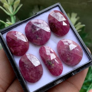 Pode incluir: Uma coleção de seis rubis facetados de formato oval, de um vermelho rosado profundo, dispostos numa caixa de exibição preta com forro interior branco. Cada pedra preciosa apresenta uma superfície polida e facetada que reflete a luz.