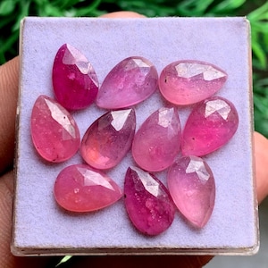May include: A collection of nine pear-shaped, faceted pink gemstones arranged on a light purple display pad. Each stone features a polished surface with geometric facets that reflect light, showcasing varying shades of vibrant pink and magenta hues.
