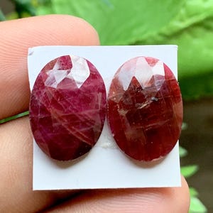 May include: Two oval-shaped, faceted rubies with a deep red colour and visible internal inclusions. The gemstones are mounted on a small white rectangular card, displaying a polished surface with geometric cuts that reflect light across the stones.