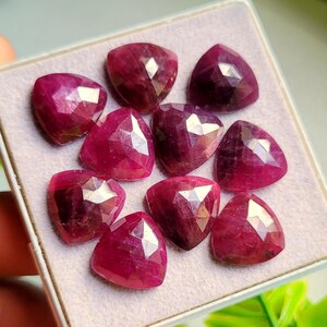 May include: A collection of nine faceted, triangular-shaped rubies with a deep magenta-red hue. Each gemstone features a polished surface with multiple geometric facets that reflect light, arranged in a grid pattern inside a square display box.