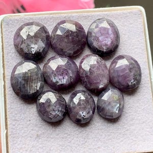 Può includere: Una collezione di dieci pietre preziose viola sfaccettate di forma ovale, disposte a griglia su un espositore in feltro grigio chiaro. Ogni pietra presenta una superficie lucida con molteplici sfaccettature geometriche che riflettono la luce.