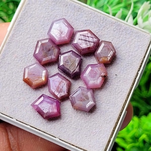 May include: A collection of nine loose, hexagonal-cut pinkish-red gemstones arranged on a light gray, textured display pad. Each stone features faceted surfaces with varying degrees of translucency, internal inclusions, and deep magenta hues.
