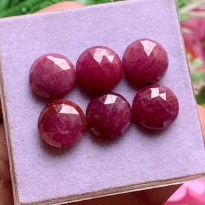 Puede incluir: Seis rubíes redondos facetados de color rojo rosado intenso, dispuestos en dos filas sobre una almohadilla de exhibición cuadrada de color violeta claro. Cada gema presenta un corte geométrico con múltiples facetas triangulares que reflejan la luz.