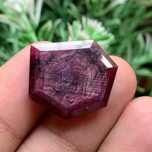 May include: A faceted, deep magenta-colored gemstone with a pentagonal shape. The stone displays a translucent quality with visible dark, linear inclusions running throughout its surface, creating a complex, textured pattern within the crystal structure.