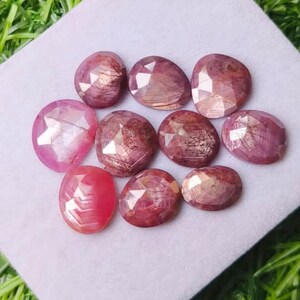 Può includere: Una collezione di dieci rubini sfaccettati di forma ovale in varie tonalità di rosa intenso e rosso. Ogni gemma presenta una superficie lucida con sfaccettature geometriche che riflettono la luce, mostrando inclusioni naturali e variazioni di colore.