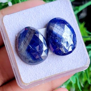 Peut inclure: Deux saphirs bleu foncé facettés en forme de poire. Les pierres précieuses sont présentées sur une surface violet clair, mettant en valeur leur taille brillante et leur couleur. Les saphirs sont d'un bleu riche et foncé, reflétant la lumière.