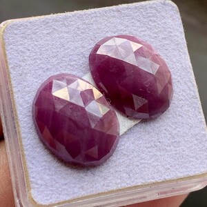 Pode incluir: Duas pedras preciosas ovais, vermelho escuro, facetadas. As gemas são apresentadas numa superfície cinza claro dentro de um recipiente de plástico transparente. As facetas capturam a luz, criando um efeito brilhante. Provavelmente rubis, para joalharia.
