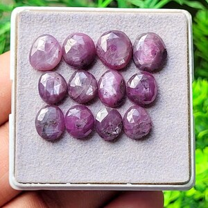 May include: A collection of eleven faceted, oval-shaped pinkish-red rubies arranged in three rows on a light gray display pad. Each gemstone features a polished, multi-faceted surface that reflects light, showcasing deep magenta and crimson hues.