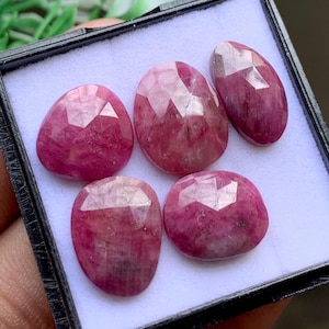Pode incluir: Cinco rubis facetados de cor rosa profundo, de formato oval, dispostos numa caixa de exposição preta com forro interior branco. Cada pedra preciosa apresenta uma superfície polida de múltiplas facetas que reflete a luz.
