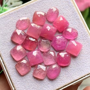 May include: A collection of loose, faceted pink gemstones in various shapes including round, oval, square, and cushion cuts. These translucent stones exhibit a vibrant pink hue and are arranged together on a light-coloured, textured display surface.