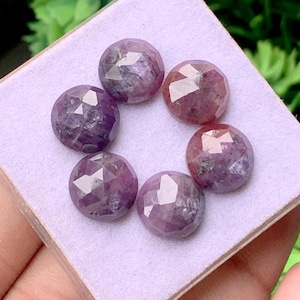 May include: Six round, faceted gemstones in shades of purple and reddish-pink. Each stone features a geometric cut with multiple flat surfaces that reflect light, arranged in a circular pattern on a light purple, square-shaped display pad.