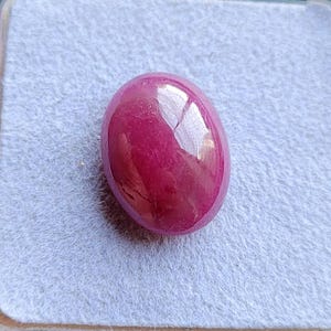 Może przedstawiać: Wypolerowany, owalny kamień szlachetny - rubin. Klejnot ma głęboki, nasycony czerwony kolor z błyszczącą powierzchnią, odbijającą światło. Rubin jest osadzony na jasnoszarym, teksturowanym tle.