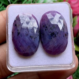 May include: Two oval-shaped, faceted purple gemstones with visible dark inclusions. The stones are set side-by-side on a white foam insert inside a clear, square plastic display case, showcasing a polished surface with geometric cut patterns.
