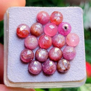 Könnte beinhalten: Eine Sammlung facettierter, runder Edelsteine in Rosa- und Rottönen, präsentiert auf einer hellgrauen Oberfläche. Die Edelsteine haben eine polierte, reflektierende Oberfläche mit unterschiedlichen Farbintensitäten. Die Anordnung ist eine Nahaufnahme.