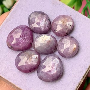 Puede incluir: Un conjunto de siete piedras preciosas facetadas de forma ovalada en tonos rosa púrpura, dispuestas en grupo. Cada piedra presenta una superficie pulida con múltiples facetas que refleja la luz, mostrando tonos translúcidos de violeta.
