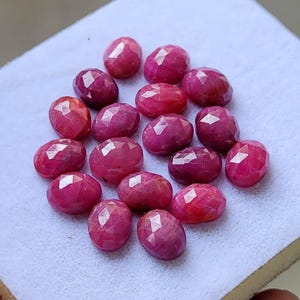 May include: A collection of fifteen oval-shaped, faceted rubies with a deep pinkish-red hue. Each gemstone features a polished surface with multiple geometric facets that reflect light, arranged in a cluster on a light-colored, textured surface.