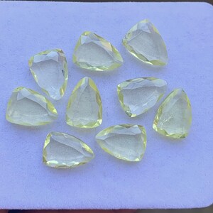 May include: Nine faceted, pale yellow gemstones with various triangular and teardrop shapes are arranged on a light surface. The gems have a translucent quality, with visible internal facets. The overall arrangement suggests a collection of loose gemstones.