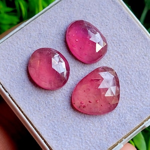May include: Three faceted, oval-shaped pink gemstones with a polished finish. The stones exhibit varying shades of deep magenta and translucent pink, featuring geometric cut patterns on their upper surfaces that reflect light across their smooth faces.