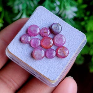 May include: A selection of polished, round gemstones in shades of pink and red, presented on a light grey surface. The gemstones vary in colour intensity, with some appearing more translucent. The arrangement is set against a blurred green background.