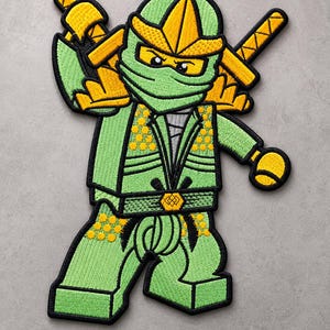 Könnte beinhalten: Ein gestickter Aufnäher, der einen grünen Ninja-Charakter mit gelber Maske und Akzenten zeigt. Die Figur trägt eine grüne Tunika mit gelben kreisförmigen Mustern, einen Gürtel mit einem zentralen gelben Emblem und zwei gelbe Schwerter auf dem Rücken.