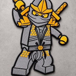 Könnte beinhalten: Ein gestickter Aufnäher, der einen grauen und gelben Ninja-Charakter mit Maske, Kapuze und zwei auf dem Rücken befestigten Schwertern zeigt. Die Figur hat ein blockartiges, gegliedertes Design mit gelben Akzenten an Brust, Beinen und Kopf.