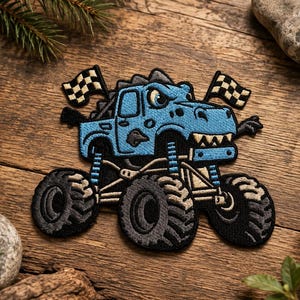 Könnte beinhalten: Ein gestickter Aufnäher, der einen hellblauen Monstertruck in Form eines Dinosaurierkopfes mit spitzen Zähnen und einem sichtbaren Auge zeigt. Der Truck hat große schwarze Reifen, blaue Federungen und zwei Zielflaggen am Heck.